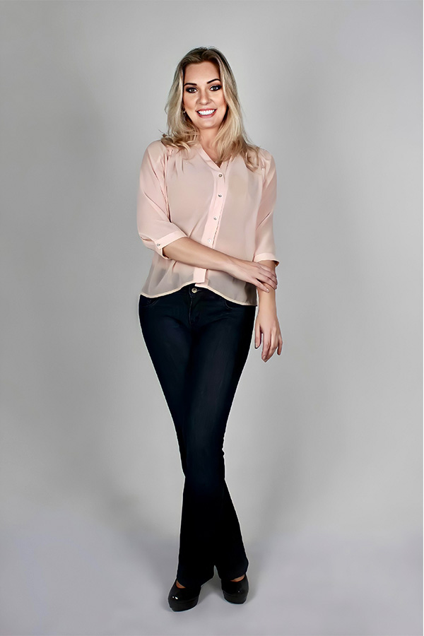 Renata Palis sorridente, em pé, vestindo camisa rosa clara e calça jeans escura, posando sobre fundo cinza.