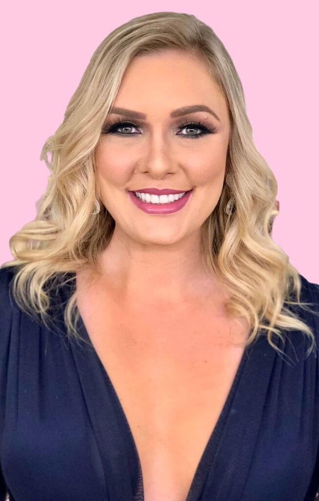 Renata Palis sorrindo, com cabelos ondulados, vestindo blusa azul com decote, posando diante de um fundo rosa claro.